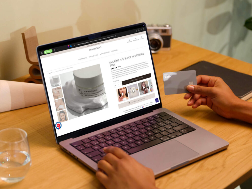 Ordinateur portable affichant la fiche produit de la crème aux « super » ingrédients 50 ml de la boutique Minimaliste, une main tenant une carte bancaire