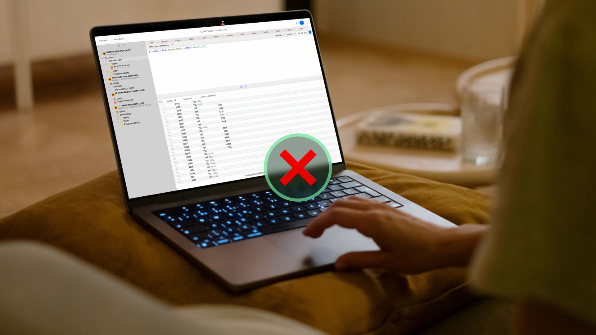 Laptop screen shows a database/table interface with a large red X overlay indicating an error or blocked access on a bed/sofa setting